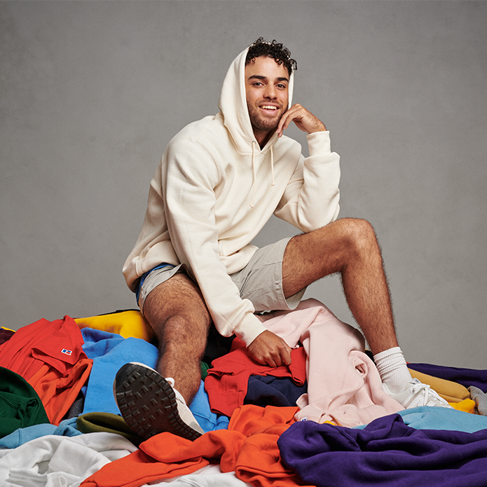 Male model wearing a cream Russell Athletic hoodie surrounded by a bunch of Russell Athletic apparel on the ground. 