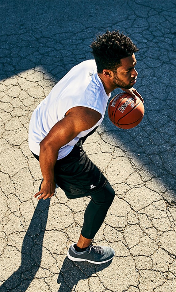 Male model dribbling a basketball in a white Russell Athletic muscle tee and black mesh shorts. 